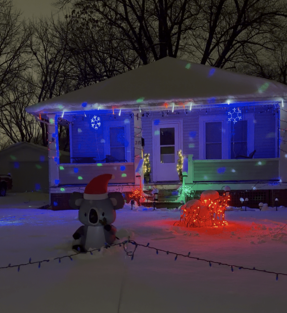 A small house is decorated with colorful holiday lights and snowflake ornaments. An inflatable koala wearing a Santa hat and illuminated reindeer sit in the snow-covered yard at night.