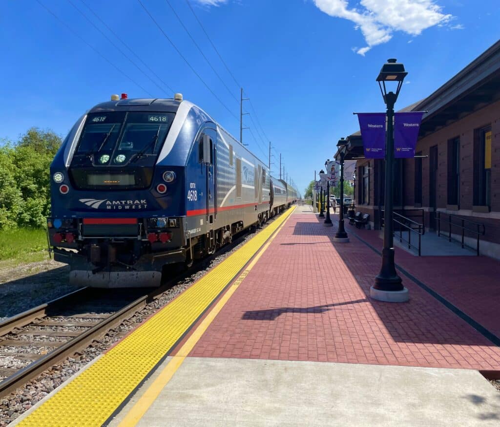 A silver and blue Amtrak train is stopped at a sunny outdoor station with a red brick platform, yellow tactile paving, and purple banners on lamp posts. The sky is clear with a few clouds.