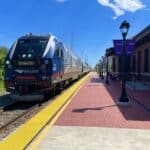 A silver and blue Amtrak train is stopped at a sunny outdoor station with a red brick platform, yellow tactile paving, and purple banners on lamp posts. The sky is clear with a few clouds.