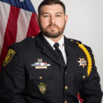 A uniformed police officer with a beard sits in front of an American flag, facing the camera. He wears a dark dress uniform with badges, pins, and a yellow braided shoulder cord.