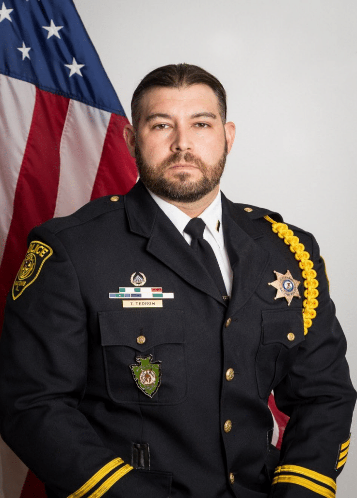 A uniformed police officer with a beard sits in front of an American flag, facing the camera. He wears a dark dress uniform with badges, pins, and a yellow braided shoulder cord.