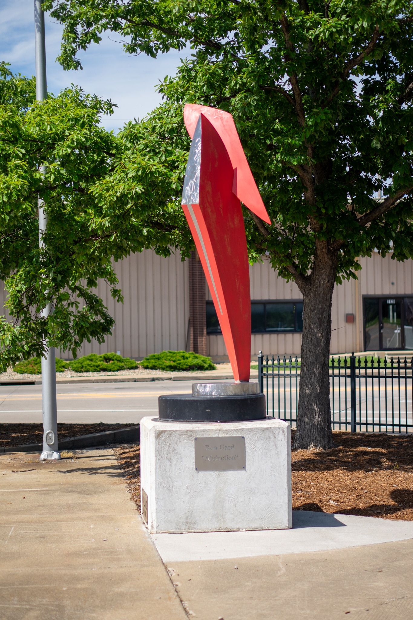 A red abstract bird sculpture stands on a white pedestal outdoors, with green trees and a building visible in the background. A plaque is mounted on the pedestal.