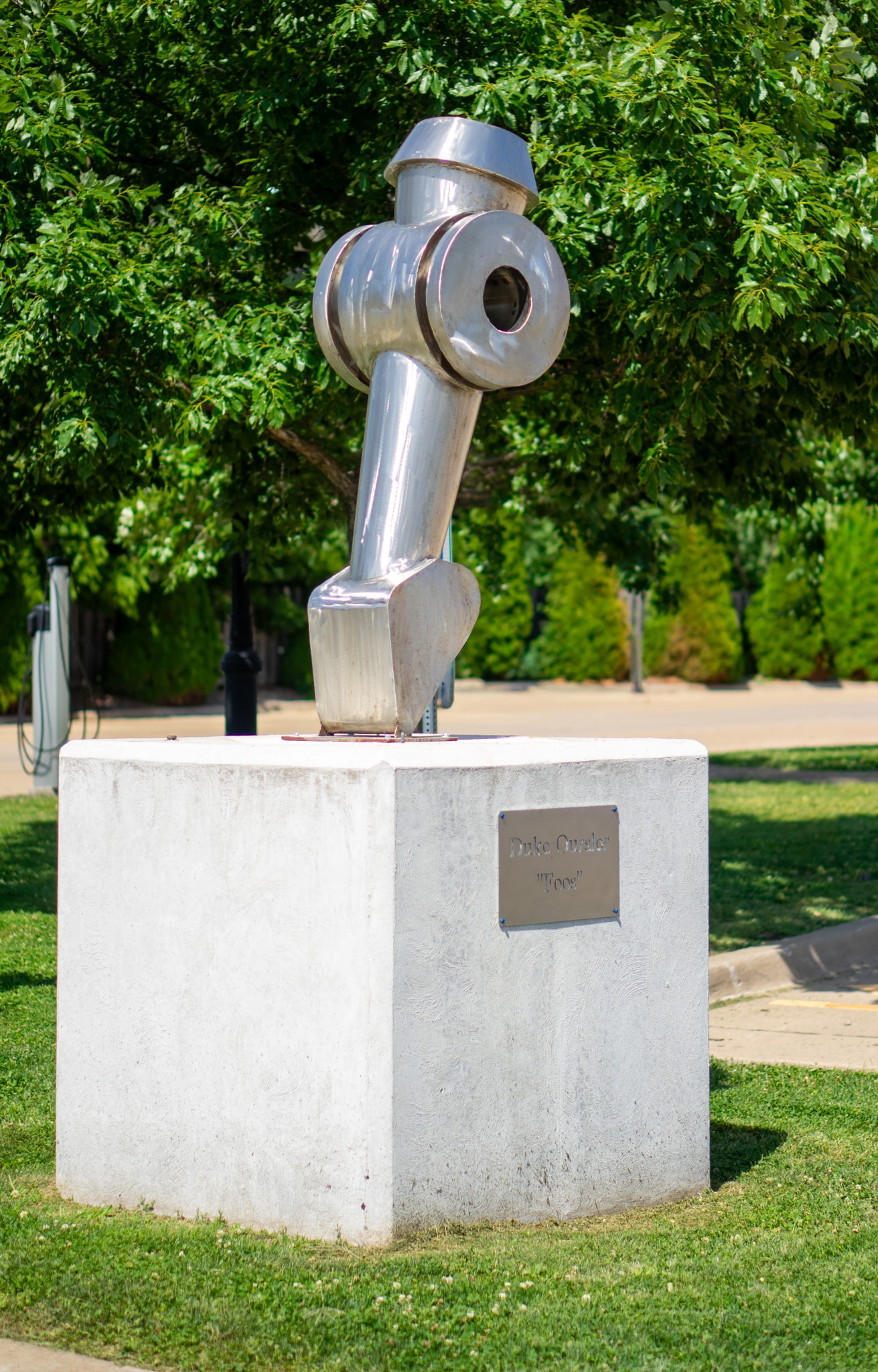 A shiny, abstract metal sculpture sits on a white concrete pedestal outdoors, surrounded by grass, trees, and sidewalks. A plaque on the pedestal displays the artist’s name and the sculpture’s title.