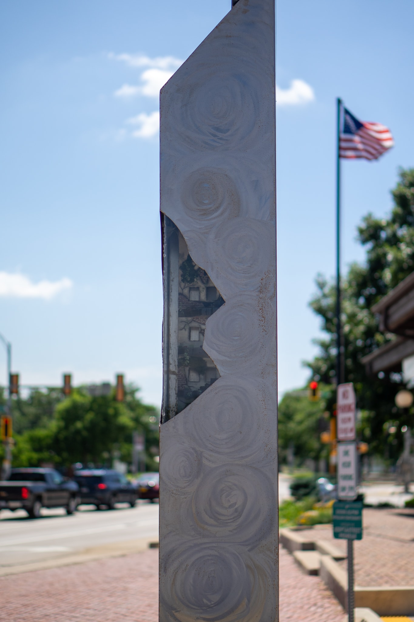 A tall, metallic sculpture with a wavy, cut-out section stands on a city sidewalk. In the background, cars drive by, trees line the street, and an American flag waves in the blue sky.