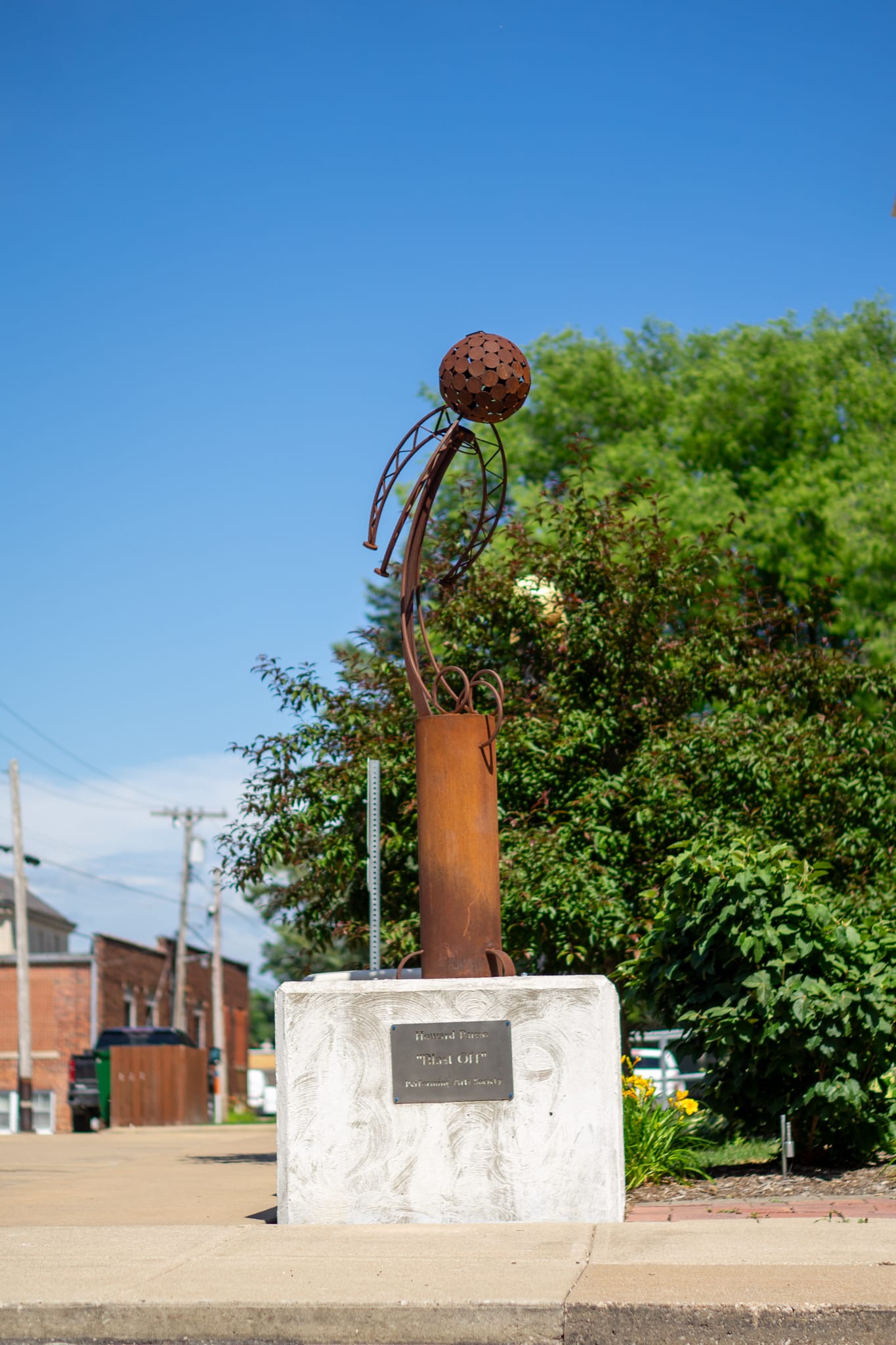 A metal sculpture featuring a spherical structure with rods is mounted on a concrete base, displayed outdoors near greenery and a sidewalk on a sunny day. A plaque is attached to the base.