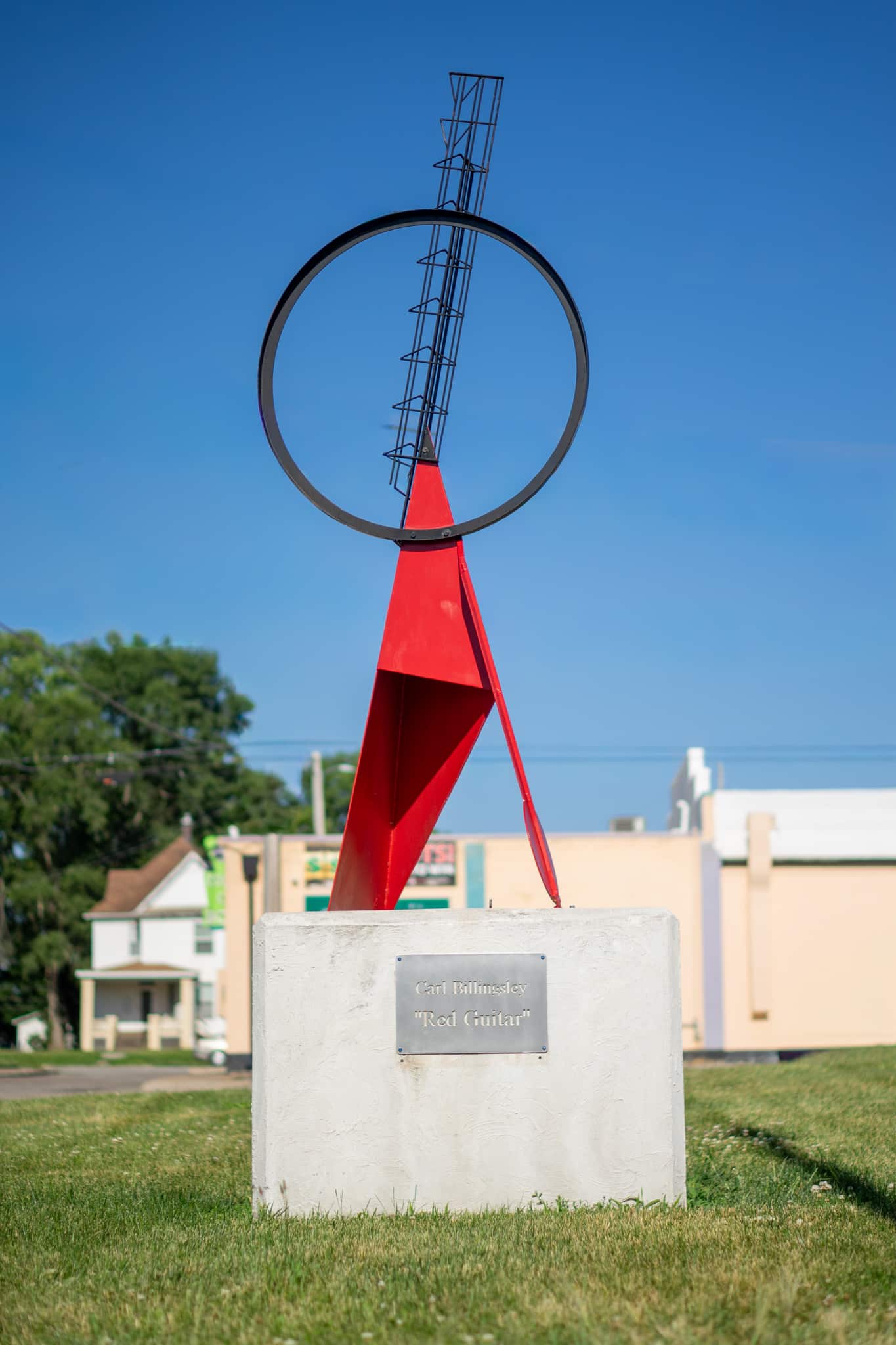 A modern outdoor sculpture with a red triangular base and a black circular frame topped by a wire ladder, displayed on a concrete platform with a plaque, set on grass with buildings and trees in the background.