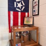 A display box with a folded flag sits beneath a hanging flag with a star and stripes. Framed informational sheets are mounted on a white brick wall beside and behind the display.