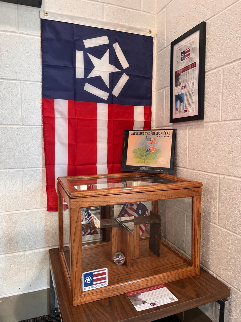 A display box with a folded flag sits beneath a hanging flag with a star and stripes. Framed informational sheets are mounted on a white brick wall beside and behind the display.