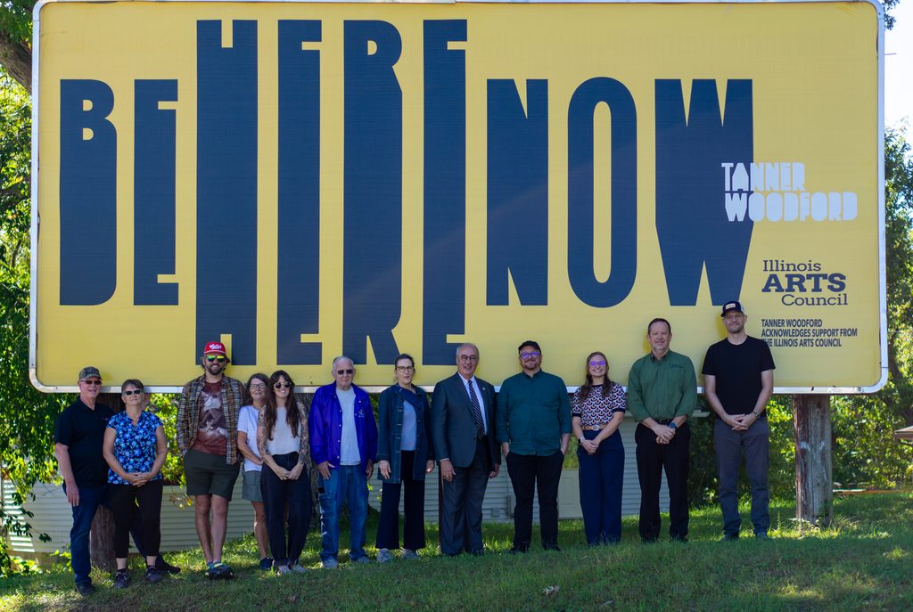 A group of eleven people standing in a row in front of a large yellow billboard that reads BE HERE NOW and TANNER WOODFORD ILLINOIS ARTS COUNCIL.