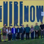 A group of thirteen people stand in front of a large yellow billboard that reads BE HERE NOW and Tanner Woodford Illinois Arts Council.