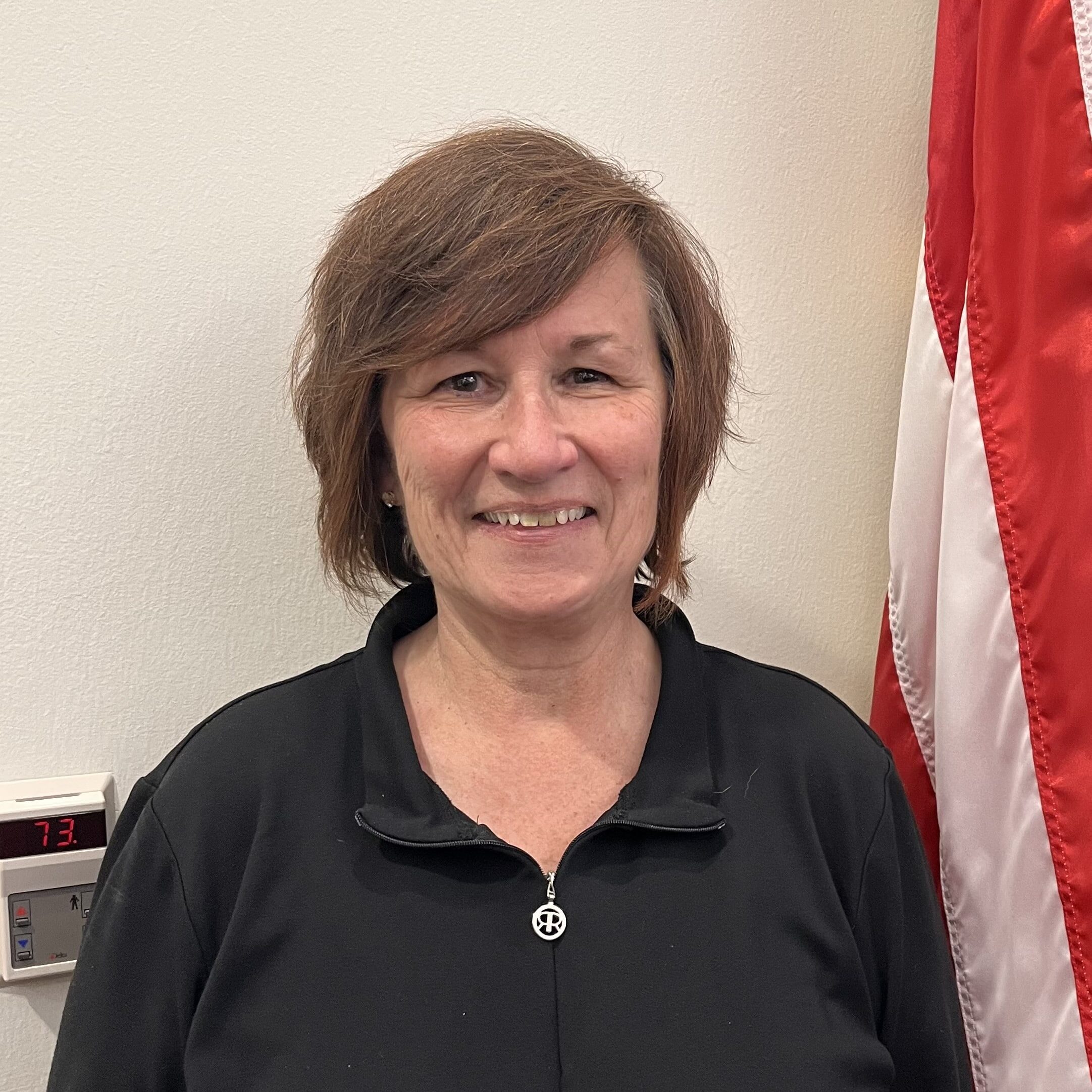 A woman with short brown hair and a black zip-up top stands and smiles in front of a white wall and a partially visible American flag.