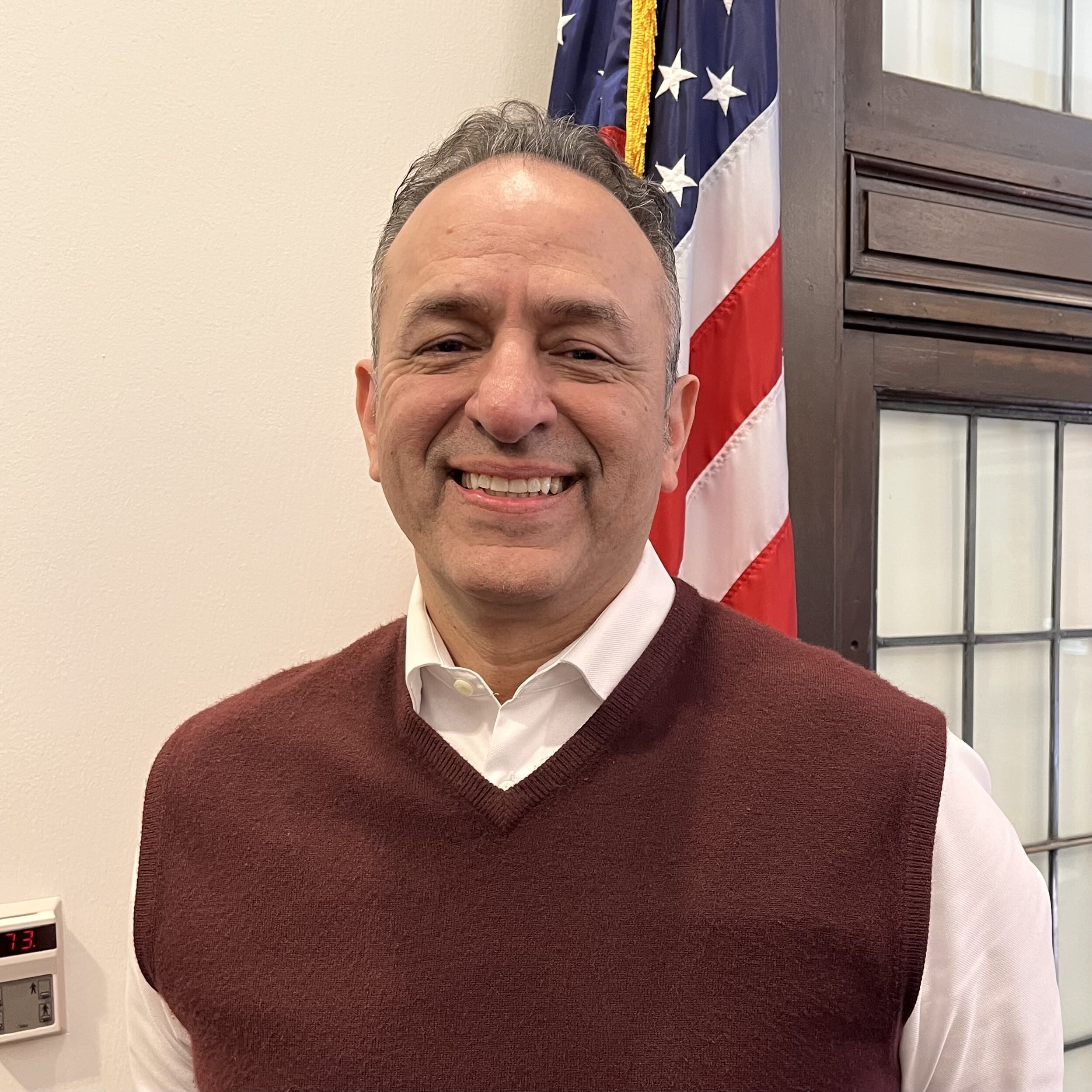 A smiling man wearing a maroon sweater vest over a white shirt stands indoors in front of an American flag and a window with wooden framing.